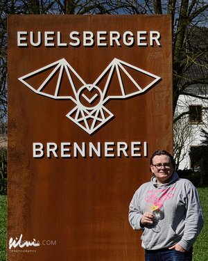 Person holding a glass standing beside a rust-colored metal sign for Euelsberger Brennerei with a bat logo.