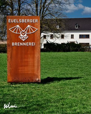 Rusty metal sign with EUELSBERGER BRENNEREI and bat logo on grass in front of white building.