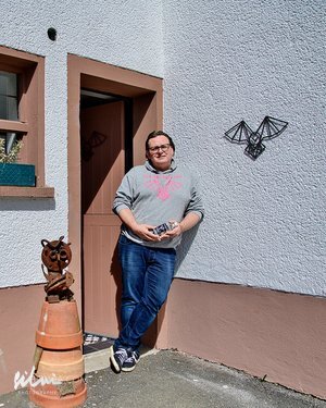 Person in glasses and casual clothes leaning against a building wall near clay pots and a metal bat decoration.