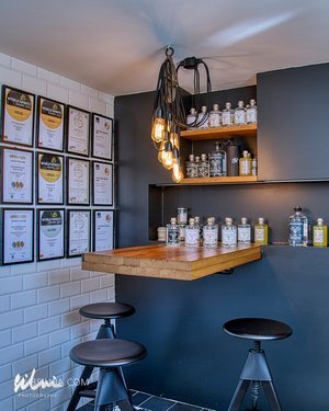 Small tasting bar with wooden counter, three black stools, hanging lights, bottles, and framed certificates.
