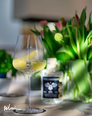 Wine glass with lemon peel on table beside a bottle and green leafy plants with pink flowers.