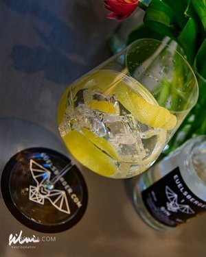 Glass with ice and lemon peels next to a branded bottle and black circular object with logo.