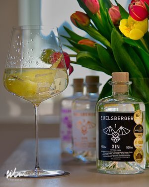 Glass with drink and ice beside Euelsberger gin bottle and tulip bouquet on surface