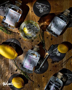 Bottles of Fuelsberger gin, lemons, black logo coasters, cocktail strainer, and a glass with ice on wood.