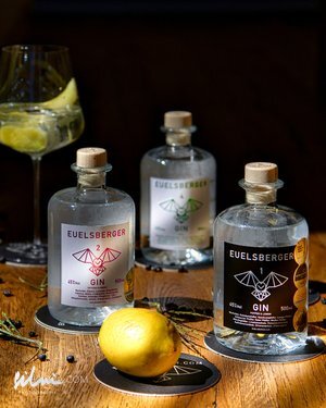 Three bottles of EUELSBERGER gin, a lemon, and a glass with lemon slices on a wooden table.