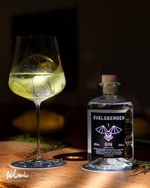 Glass of gin with ice and lemon next to a clear Euelsberger Gin bottle on wood surface.