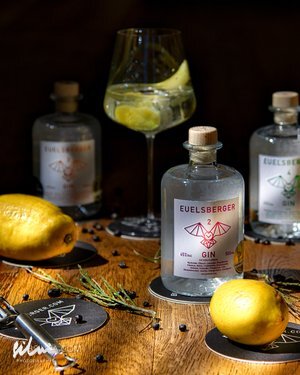 Bottles of Euelsberger gin, lemons, juniper berries, rosemary, and a gin and tonic glass on wood.