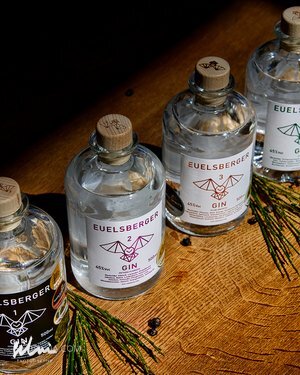 Four bottles of EUELSBERGER gin with wooden caps on a wooden surface with pine sprigs and berries.