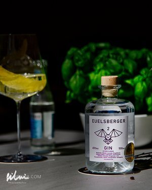 Bottle of Euelsberger Gin with wooden cork beside a glass with lemon peel and water bottle on table