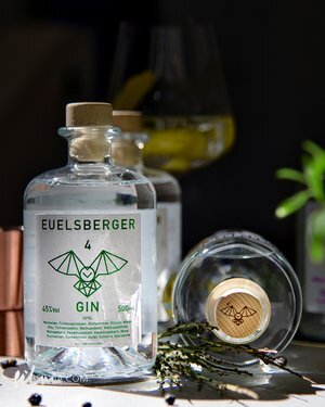 Clear EUELSBERGER gin bottles with cork stoppers, herbs, and a glass with a yellow drink on dark surface.