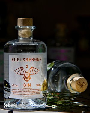 Clear Euelsberger Gin bottle with wooden cork, bat label, 45% alcohol, 500ml, herbs nearby.
