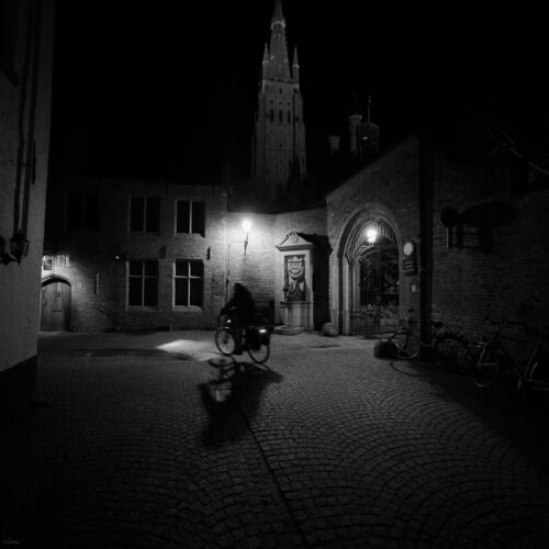 Nighttime black and white scene of a cyclist in a cobblestone courtyard with brick buildings and a tower.
