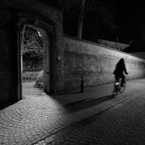 Person riding a bicycle on a cobblestone path beside a lit arched doorway in a brick wall at night.