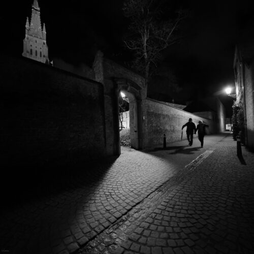 Nighttime cobblestone street with two people walking, one holding a cane, near an archway.