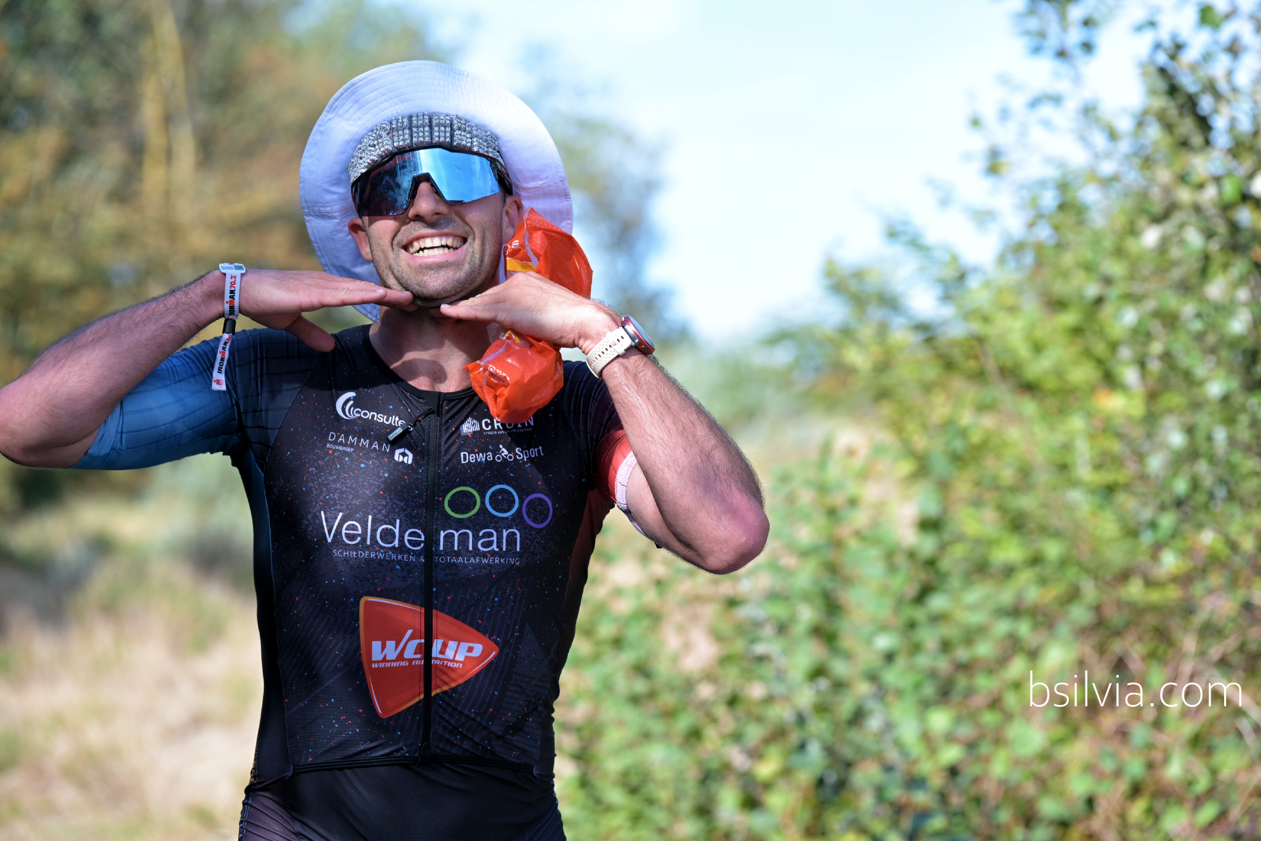 Smiling IRONMAN Knokke 2026 participant wearing mirrored sunglasses and a white hat, posing with both hands beneath his chin.